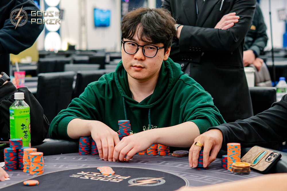 Man with glasses and green hoodie sitting at a poker table with chips, looking focused.