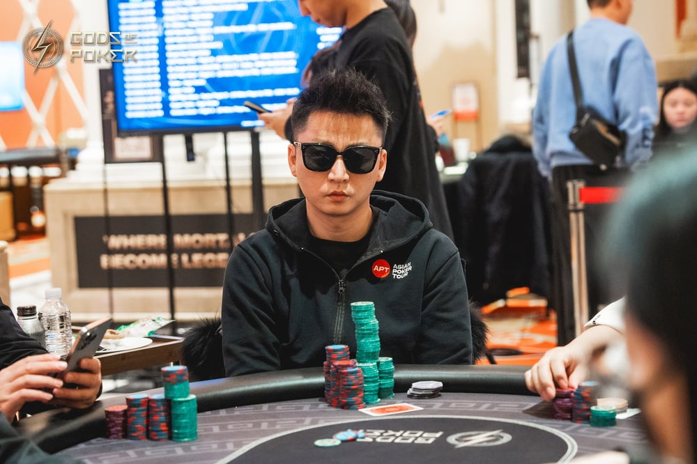 A man in sunglasses and a black hoodie sits at a poker table with chips.