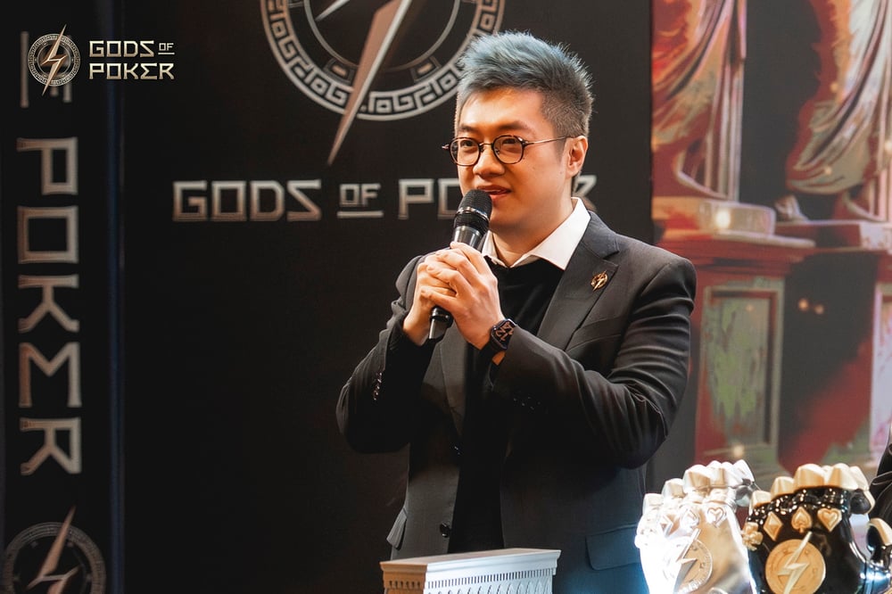 A man in a suit and glasses speaks into a microphone at a 'Gods of Poker' event.