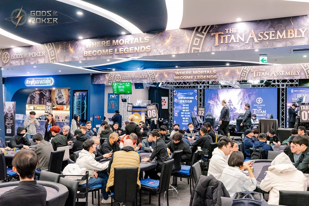 Busy poker tournament room with many players at tables under 'Gods of Poker' banners.